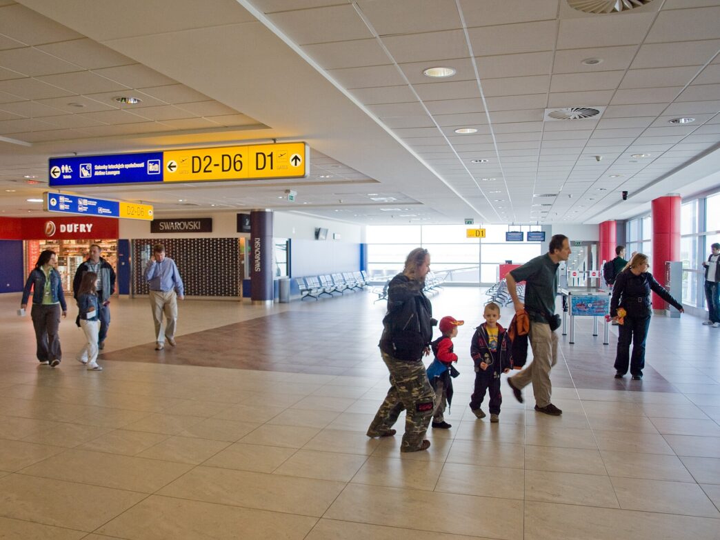 Presvetlená chodba terminálu so žltými smerovkami k východom D1 a D2, chodbou kráča niekoľko cestujúcich vrátane detí.