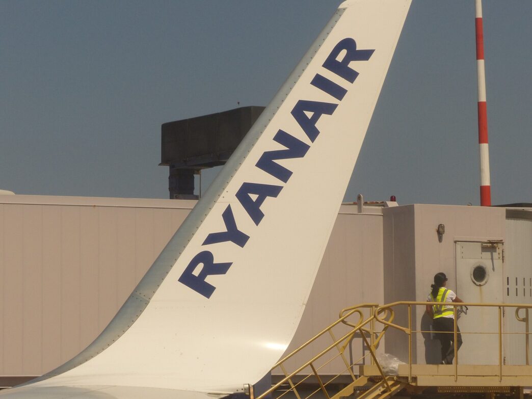 Modrý chvost lietadla Ryanairu s bielym nápisom, pod ním na plošine stojí pracovník v reflexnej veste, nad tým modrá obloha.