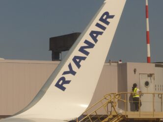 Modrý chvost lietadla Ryanairu s bielym nápisom, pod ním na plošine stojí pracovník v reflexnej veste, nad tým modrá obloha.