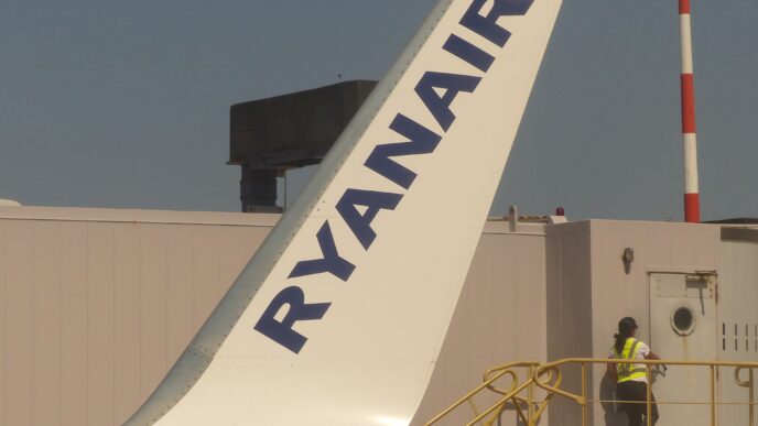 Modrý chvost lietadla Ryanairu s bielym nápisom, pod ním na plošine stojí pracovník v reflexnej veste, nad tým modrá obloha.