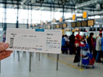 Ruka drží papierový palubný lístok v odletovej hale letiska, v pozadí cestujúci s batožinou a informačné tabule