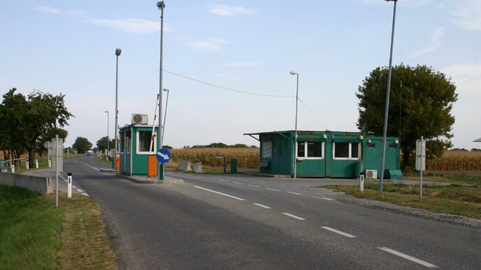 Cestná hraničná stanica so zdvihnutou rampou a zelenými kontrolnými búdkami pri ceste medzi poliami