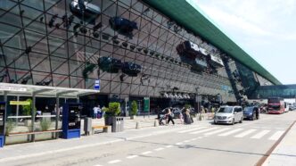 Presklená fasáda terminálu letiska Krakov, pred ňou autobusová zastávka s automatom na lístky, zaparkované autá pri kraji vozovky, cestujúci s kuframi prechádzajú po priechode a vpravo stojí zájazdový autobus.