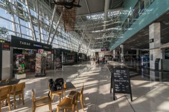 Obchodná zóna v hale terminálu s výkladmi predajní a kaviarenskými stolíkmi, popri nich prechádzajú cestujúci, cez presklenú stenu svieti denné svetlo.