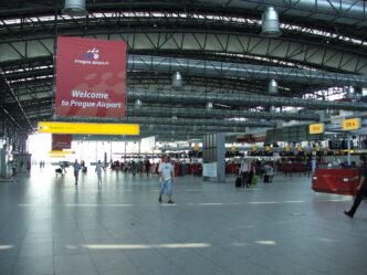 Presvetlená odbavovacia hala letiska s radom odbavovacích priehradiek, cestujúci s batožinou prechádzajú halou.
