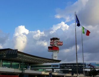 Riadiaca veža letovej prevádzky na letisku Rím-Fiumicino, vedľa nej vlajky Európskej únie a Talianska, na oblohe kopovité oblaky