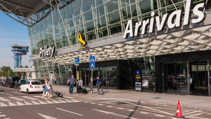Presklený vchod do príletovej haly s veľkým nápisom Prílety, pred ním stojí niekoľko ľudí s batožinou a zaparkované auto.