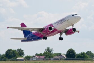 Bielo-purpurové lietadlo Airbus A320 spoločnosti Wizz Air stúpa krátko po vzlete, v pozadí zelené polia a obloha s oblakmi.