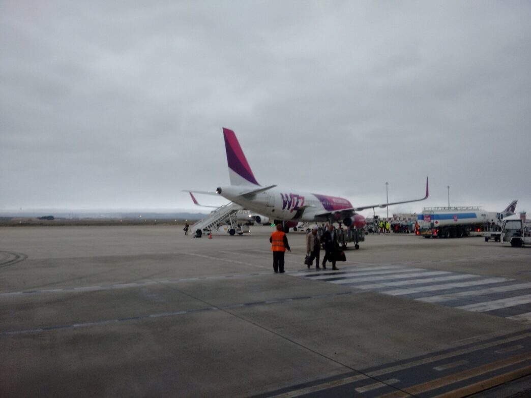 Bielo-purpurové lietadlo Wizz Air stojí na odbavovacej ploche pod zamračenou oblohou, pri ňom skupinka cestujúcich a pracovník v oranžovej veste.
