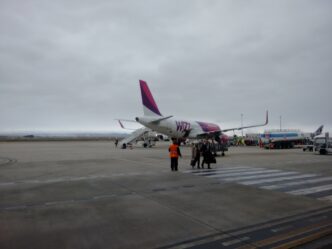 Bielo-purpurové lietadlo Wizz Air stojí na odbavovacej ploche pod zamračenou oblohou, pri ňom skupinka cestujúcich a pracovník v oranžovej veste.