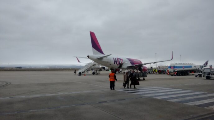 Bielo-purpurové lietadlo Wizz Air stojí na odbavovacej ploche pod zamračenou oblohou, pri ňom skupinka cestujúcich a pracovník v oranžovej veste.