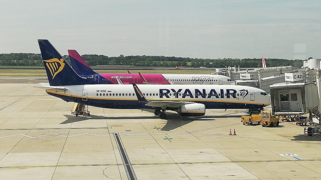 Lietadlo Ryanair v modro-bielom nátere a za ním lietadlo Wizz Air v ružovo-bielom nátere na odbavovacej ploche pri termináli, v popredí pozemná technika