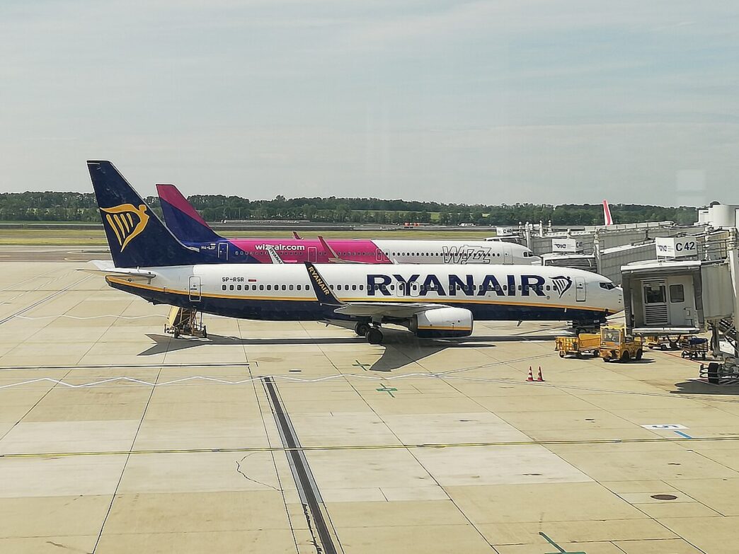 Lietadlo Ryanair v modro-bielom nátere a za ním lietadlo Wizz Air v ružovo-bielom nátere na odbavovacej ploche pri termináli, v popredí pozemná technika