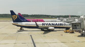 Lietadlo Ryanair v modro-bielom nátere a za ním lietadlo Wizz Air v ružovo-bielom nátere na odbavovacej ploche pri termináli, v popredí pozemná technika