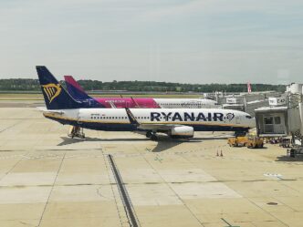 Lietadlo Ryanair v modro-bielom nátere a za ním lietadlo Wizz Air v ružovo-bielom nátere na odbavovacej ploche pri termináli, v popredí pozemná technika