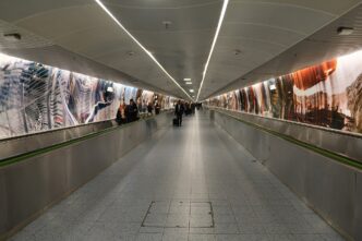Dlhá presvetlená podzemná chodba s pohyblivými chodníkmi a veľkoplošnými fotografiami na stenách, ktorou prechádzajú cestujúci s kuframi
