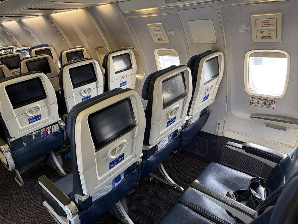 Kabína lietadla s radmi sedadiel pri dvoch núdzových východoch nad krídlom, pri oknách nálepky s pokynmi