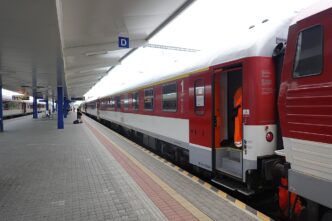 Diaľkový vlak s červeno-bielymi vozňami pri zastrešenom nástupišti železničnej stanice, v diaľke cestujúci